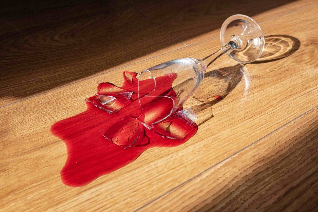 Ein zerbrochenes Weinglas auf einem Parkettboden. Der Wein läuft über die Lackschutzschicht des Parketts.