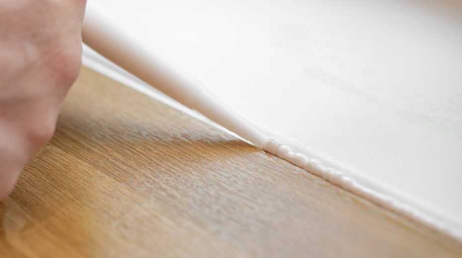 Weißes Silikon wird am Übergang zwischen einer weißen Wand und einem Holzboden angebracht.