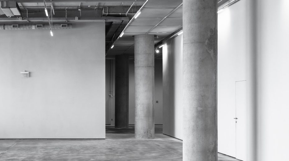 Eine Halle mit Schattenfugen zwischen den weißen Wänden und dem grauen Boden.